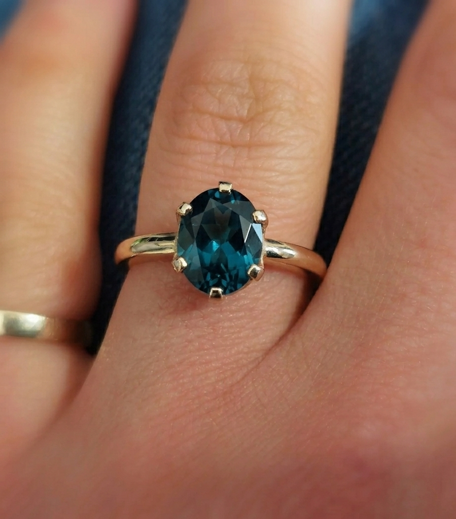 Close-up photograph of an oval-cut deep teal London blue topaz solitaire ring set in a polished yellow gold band